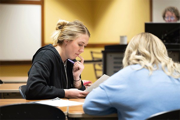 Blonde female student looking over a paper in class with another student whose back is to the camera and a professor visible in the background.