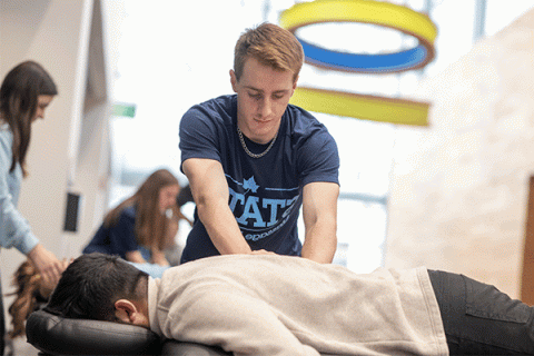 A male student with light brown hair, wearing a slate-blue t-shirt that says STATE on the front, presses down with both hands to massage the back of a man lying facedown on a massage table. The man on the table has short, dark hair and wears grey jeans and a beige shirt. In the background, slightly out of focus, two female students can be seen working with clients on other tables.   